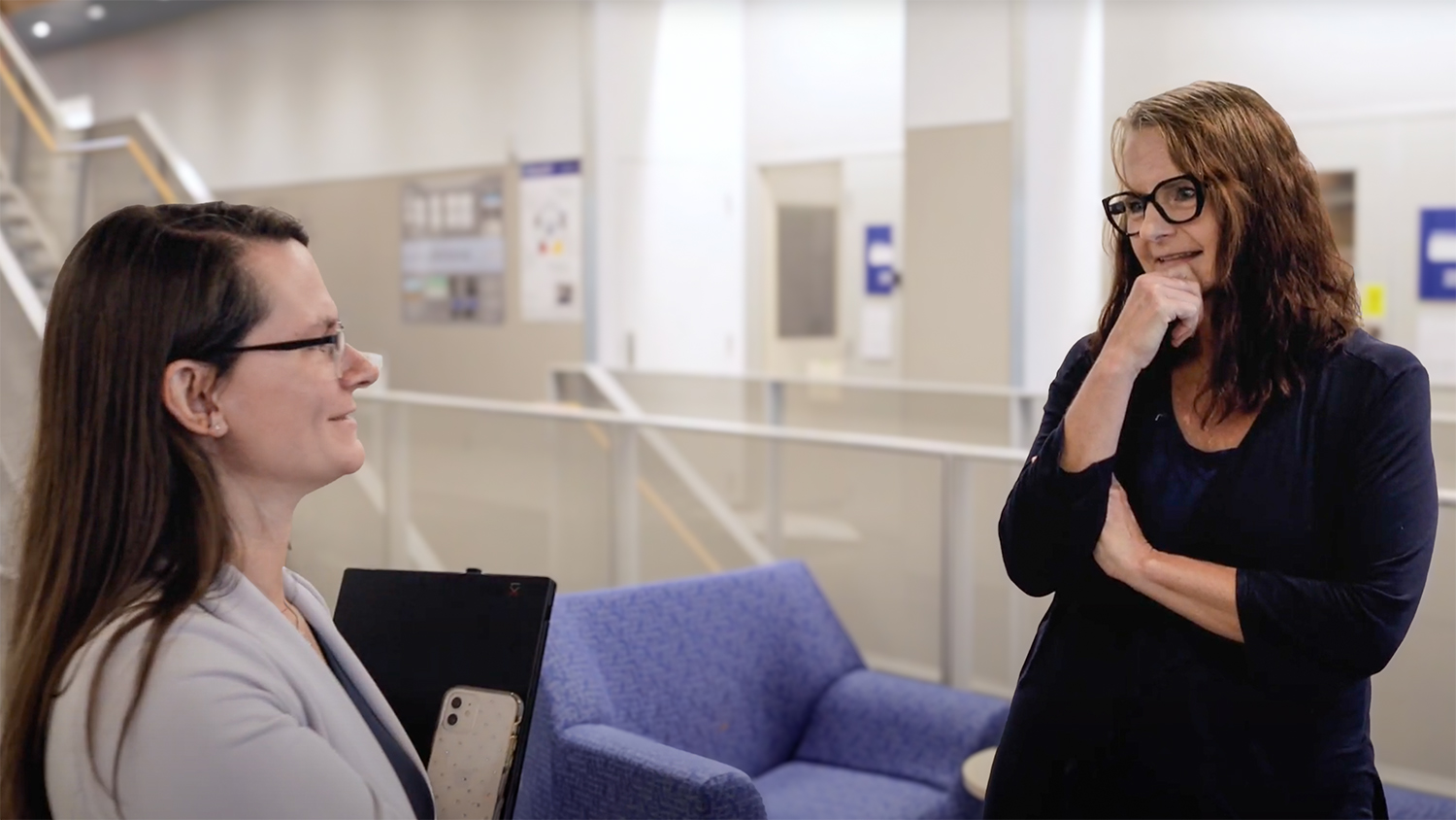 Two women with long brown hair talking inside a building with a white interior, standing next to a purple chair.